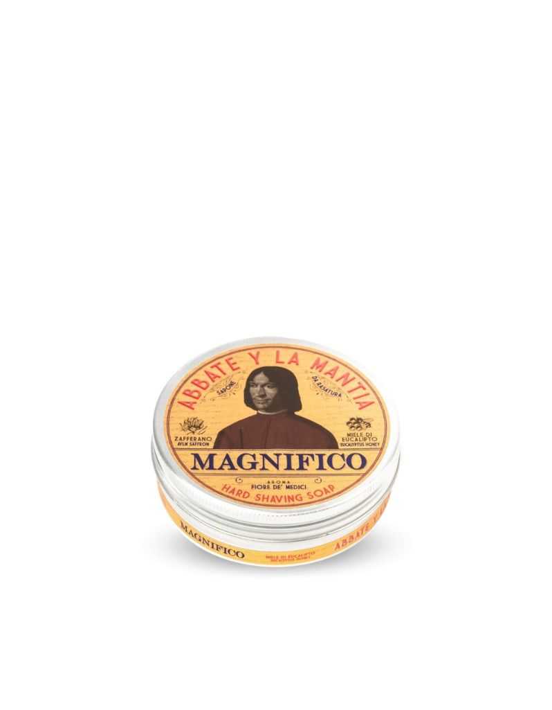 A solid shaving soap puck packaged in a round container with a lid featuring text and a portrait illustration. The label reads 'MAGNIFICO' and includes the words 'Solid Shaving Soap' as well as a list of ingredients like Eucalyptus Honey, Saffron.