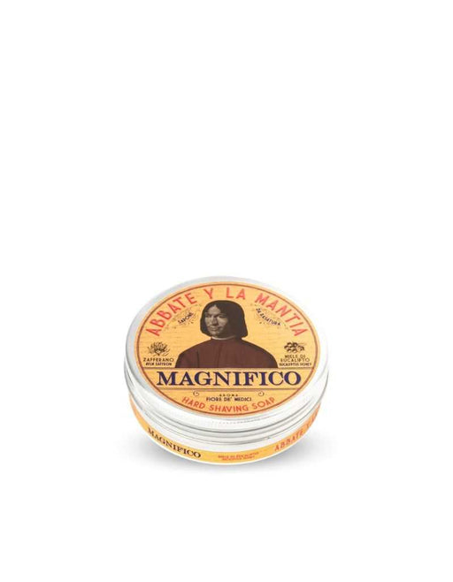 A solid shaving soap puck packaged in a round container with a lid featuring text and a portrait illustration. The label reads 'MAGNIFICO' and includes the words 'Solid Shaving Soap' as well as a list of ingredients like Eucalyptus Honey, Saffron.