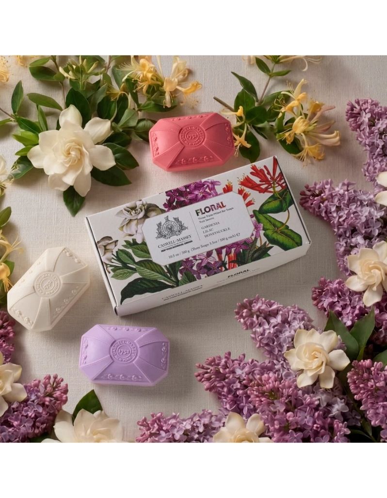 Three hexagonal bar soaps in pink, white, and lavender are arranged around a floral illustrated box labeled FLORAL, styled with white and yellow flowers on a light fabric background.