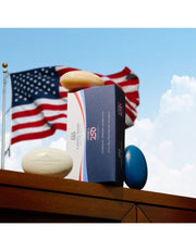 A box of America250 soap bars displayed on a wooden ledge outdoors, with an American flag and blue sky in the background. The box is navy blue with white and red accents, and three soap bars in beige, white, and blue are arranged around it.