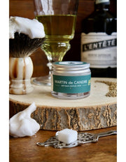 A small jar of Absinthe Martin de Cendre artisanal shaving soap sits on a rustic wood slice, surrounded by shaving props including a shaving brush with lather, a glass of olive oil, and a dark bottle in the background.