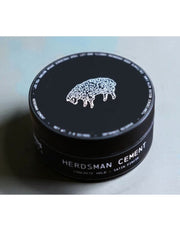 A black round tin of Herdsman Cement hair product with a white sheep logo and white text, photographed on a light gray surface.
