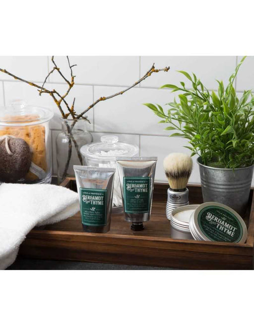 A shaving set including a boar bristle brush with an aluminum handle, tubes of shave cream and after shave balm, a tin container, and a plant in the background.