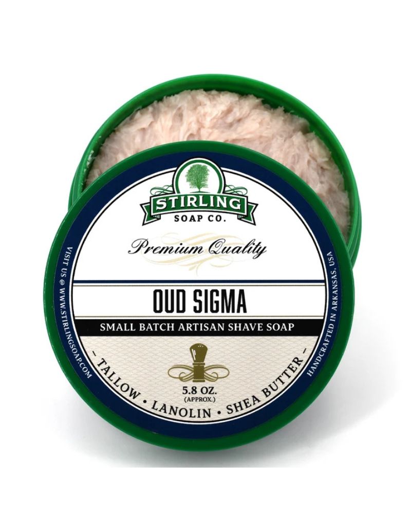 An open round tub of Stirling Soap Co. Oud Sigma artisan shave soap, showing the pale pinkish-beige soap inside with a green-rimmed container and a white label.