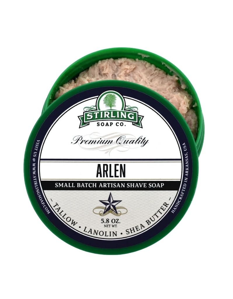 An open round tub of Stirling Soap Co. Arlen shave soap, showing the pale beige lather inside a dark green container with a white and green label.
